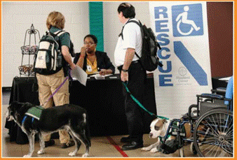 Two people with assistance dogs stand in front of a table. A woman sits behind the table and gives them papers. There is a sign on that wall that says "RESCUE."